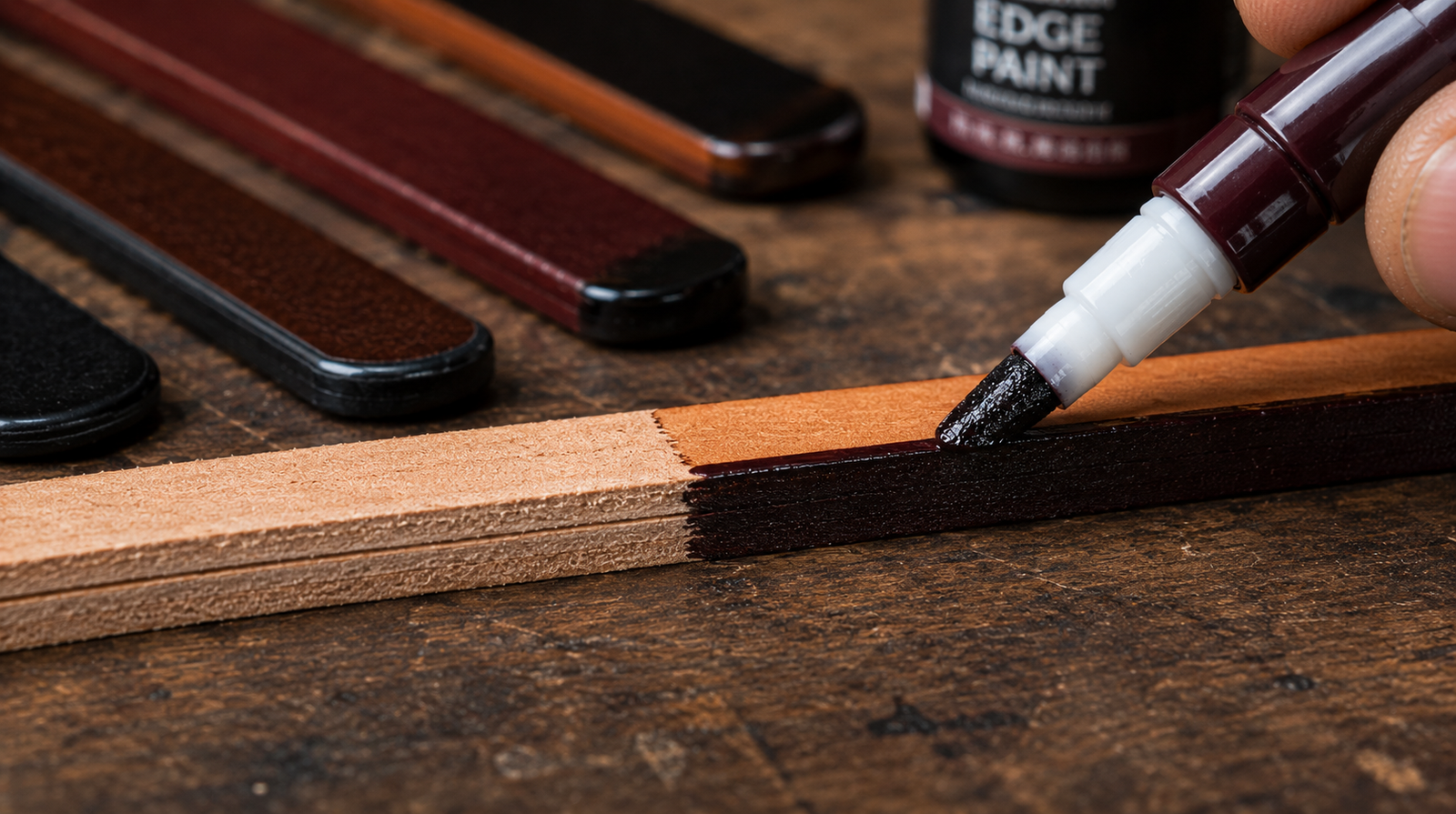 A roller pen applicator laying a thin coat of dark burgundy edge paint onto the cut edge of a natural leather strap, with finished sample strips in black, burgundy, and brown visible in the background alongside an edge paint bottle